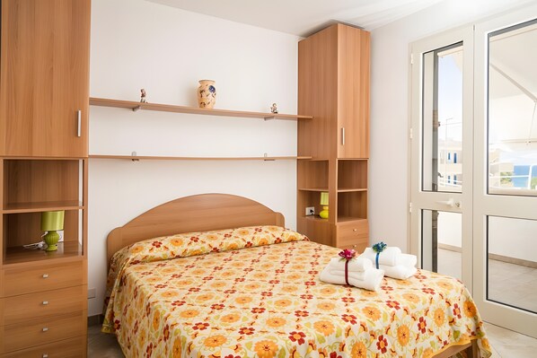 A bedroom with a wooden bed, a nightstand, a shelf with decorative items, and a window with a view.