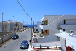 A residential street with modern buildings, a parked car, and a clear view of the sea.