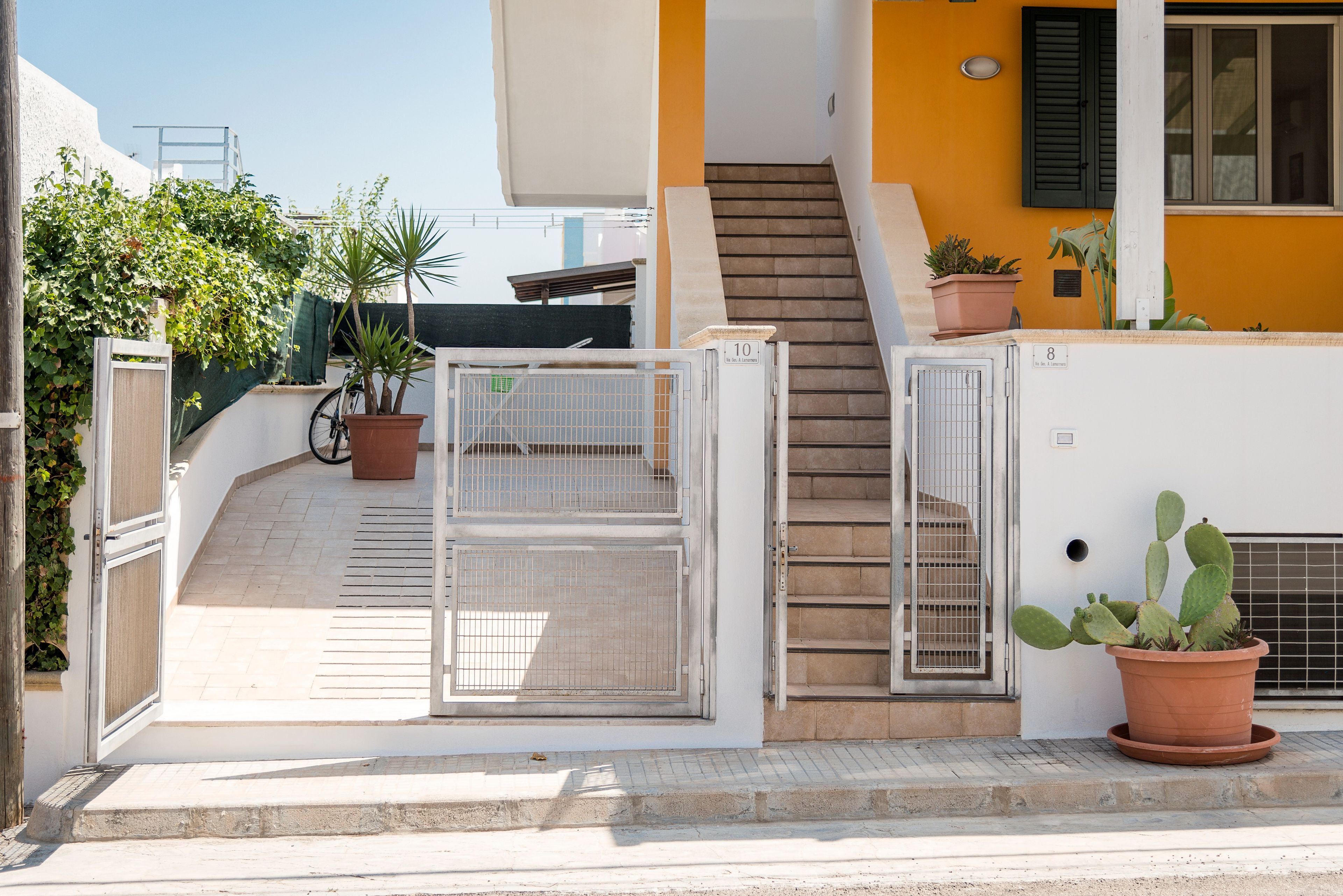 Un edificio residencial con entrada protegida por un portón, una escalera que conduce a un piso superior y plantas en macetas.