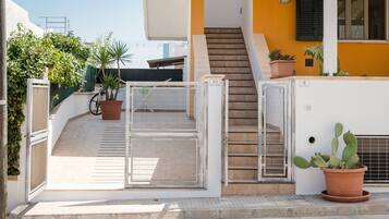 Un edificio residencial con entrada protegida por un portón, una escalera que conduce a un piso superior y plantas en macetas.