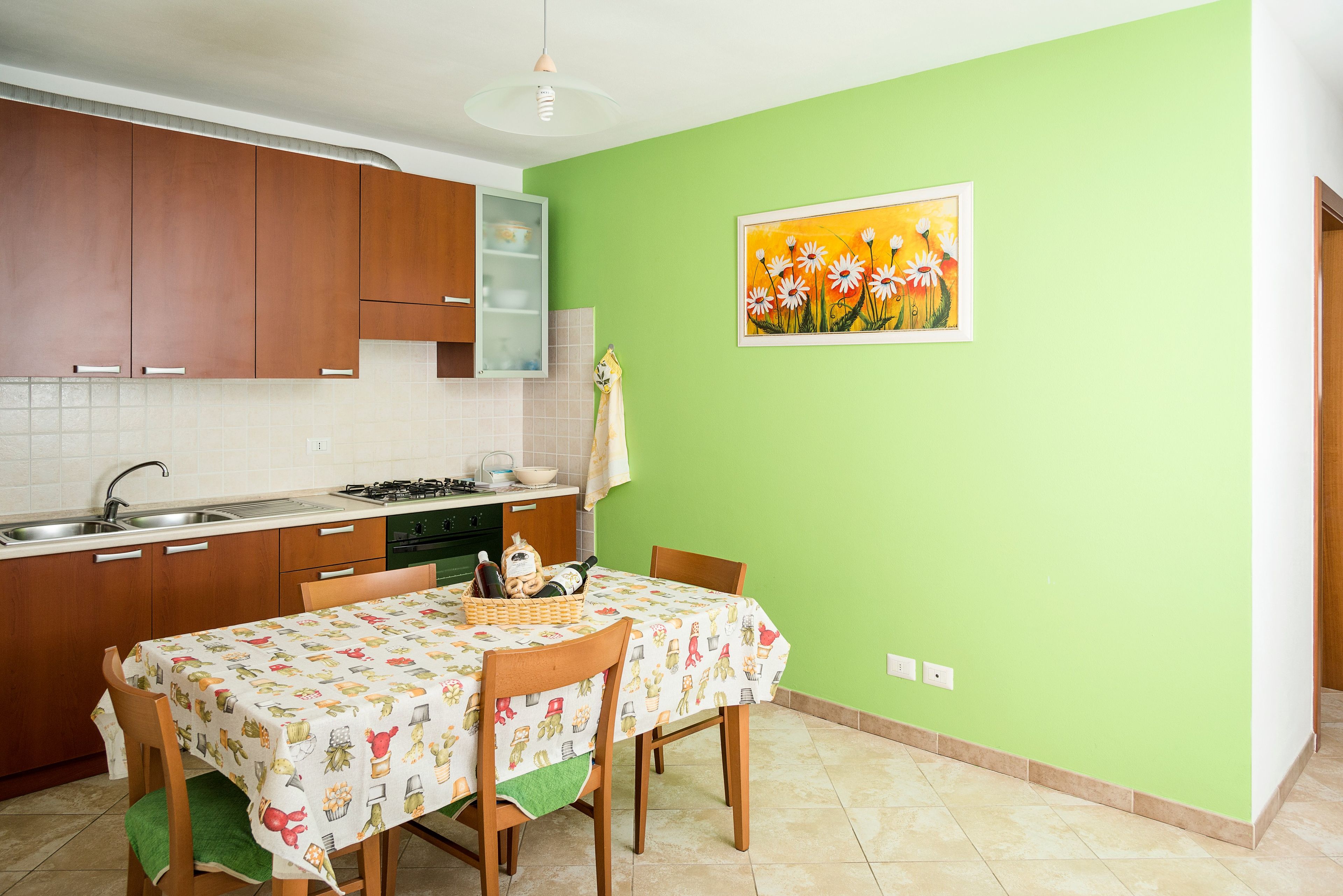 Une cuisine avec des meubles en bois, une table à manger recouverte d’une nappe à motifs floraux et un mur vert orné d’un tableau encadré.