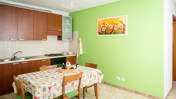 Une cuisine avec des meubles en bois, une table à manger recouverte d’une nappe à motifs floraux et un mur vert orné d’un tableau encadré.