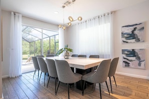 Een moderne eetruimte met een grote tafel, gestoffeerde grijze stoelen en een potplant.