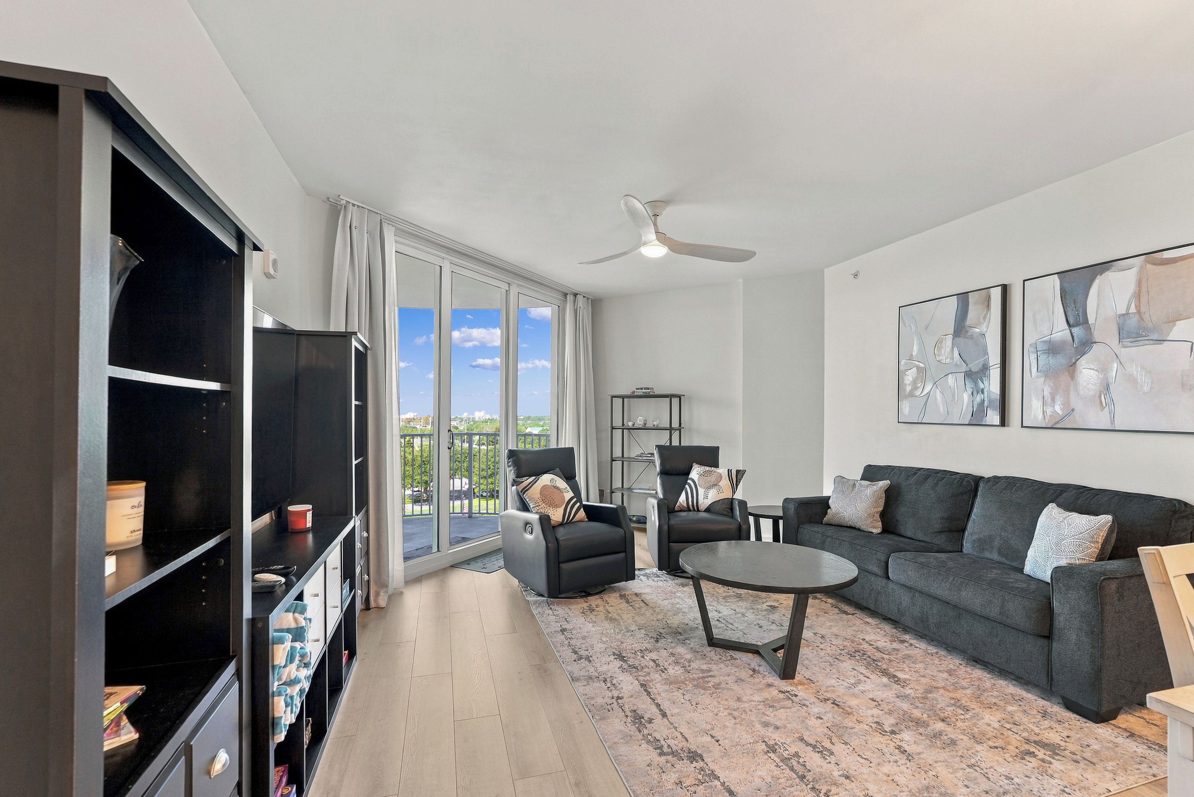 A modern living room with a large window, a ceiling fan, a black leather sofa, a coffee table, and a bookshelf.