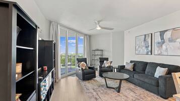 A modern living room with a large window, a ceiling fan, a black leather sofa, a coffee table, and a bookshelf.