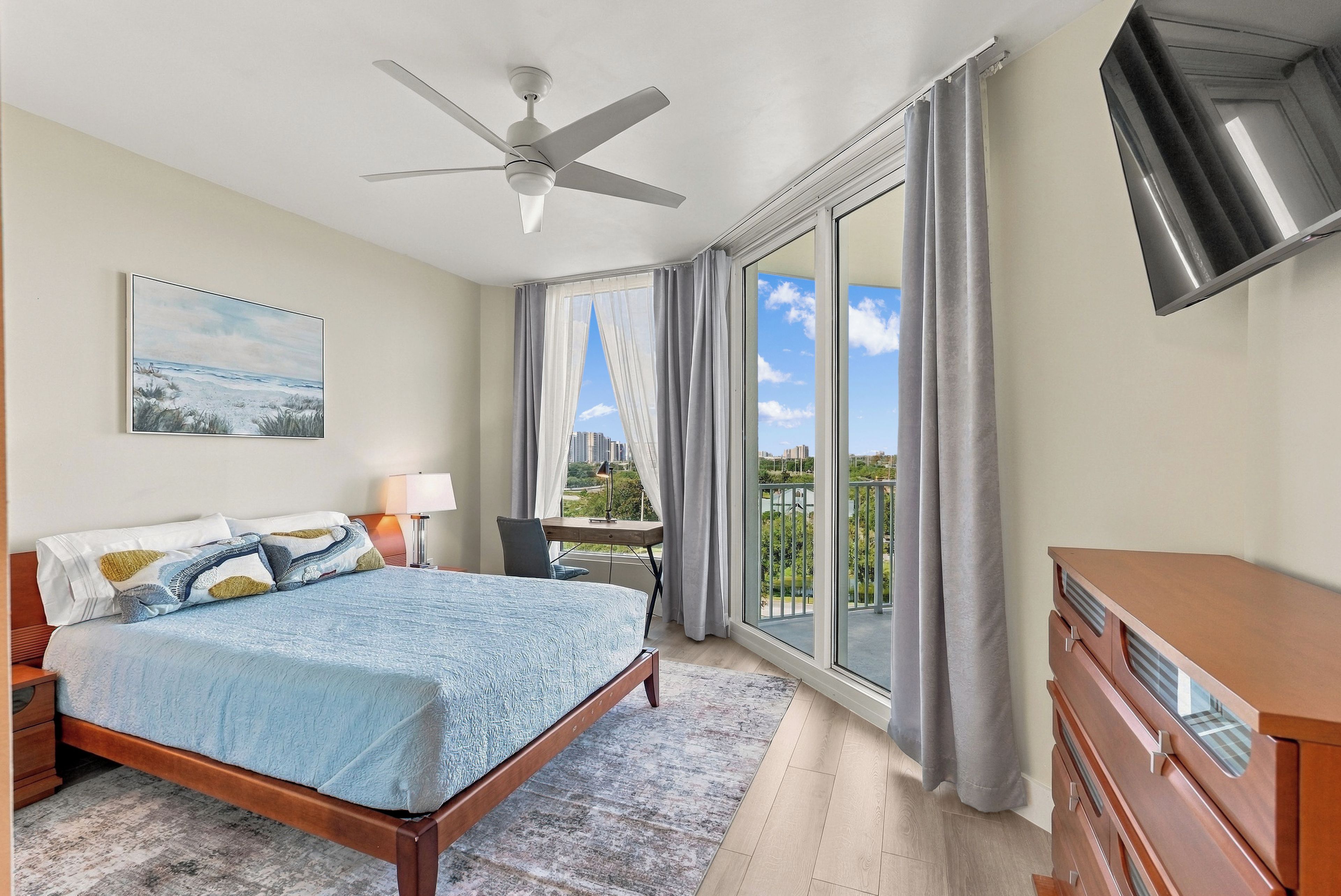 A bedroom with a bed, a desk, a chair, a TV, and a balcony with a view of buildings.