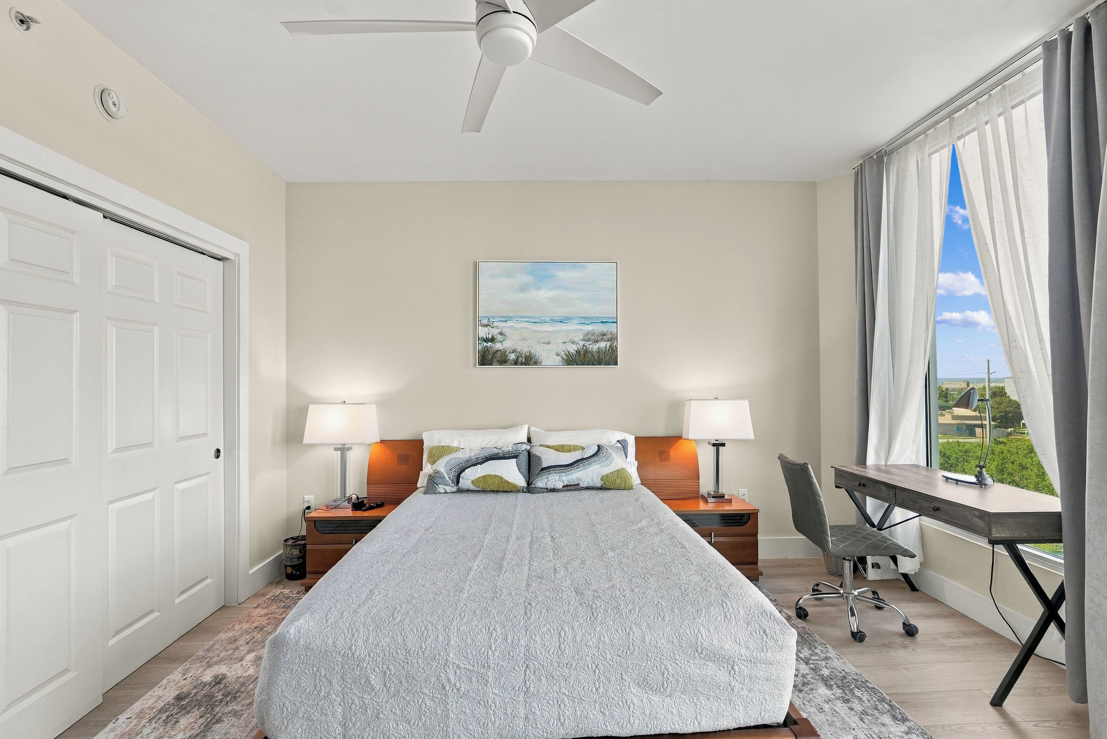 A bedroom with a bed, two bedside tables with lamps, a desk with a chair, a ceiling fan, a painting on the wall, and a window with curtains.
