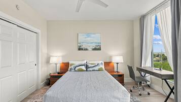 A bedroom with a bed, two bedside tables with lamps, a desk with a chair, a ceiling fan, a painting on the wall, and a window with curtains.