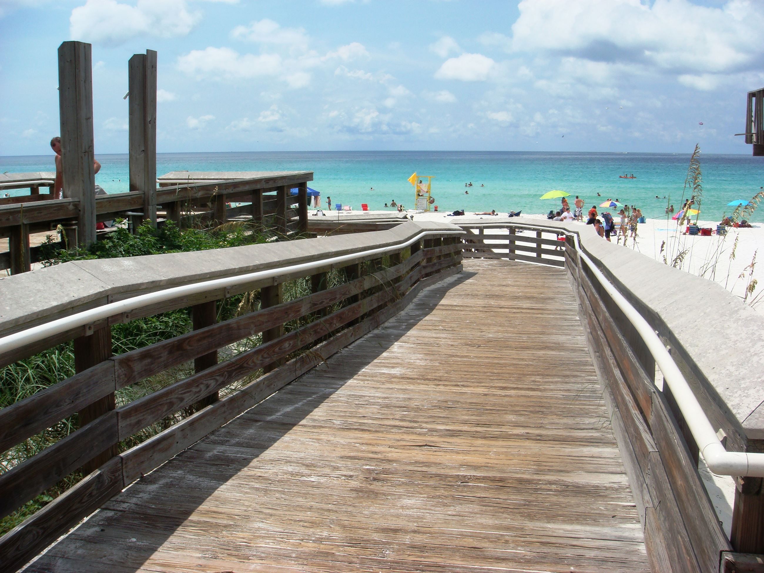 A wooden boardwalk leading to a beach with clear turquoise water and a sandy shore.