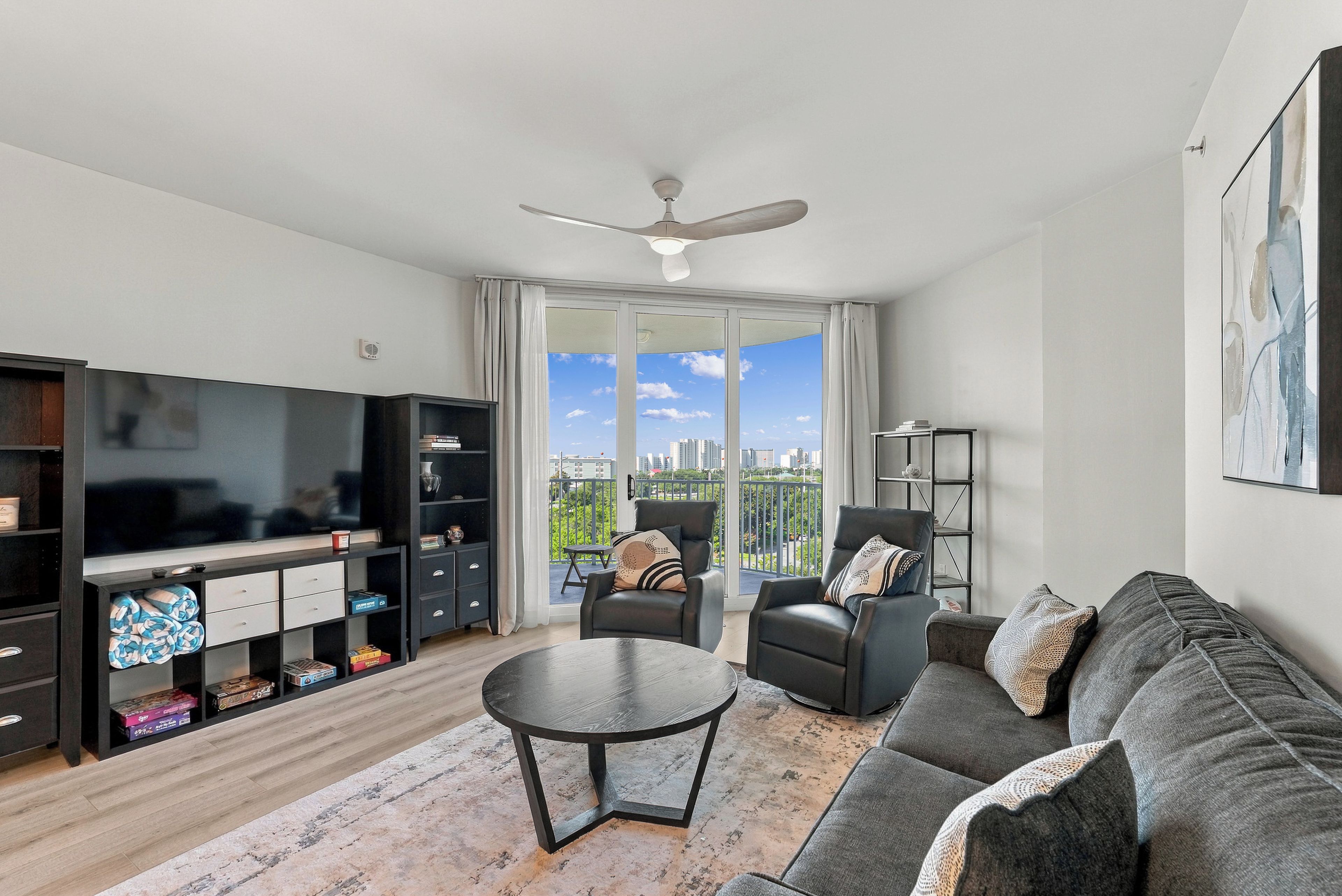 A modern living room with a flat-screen TV, a ceiling fan, a sofa, a coffee table, and a view of a cityscape through large windows.