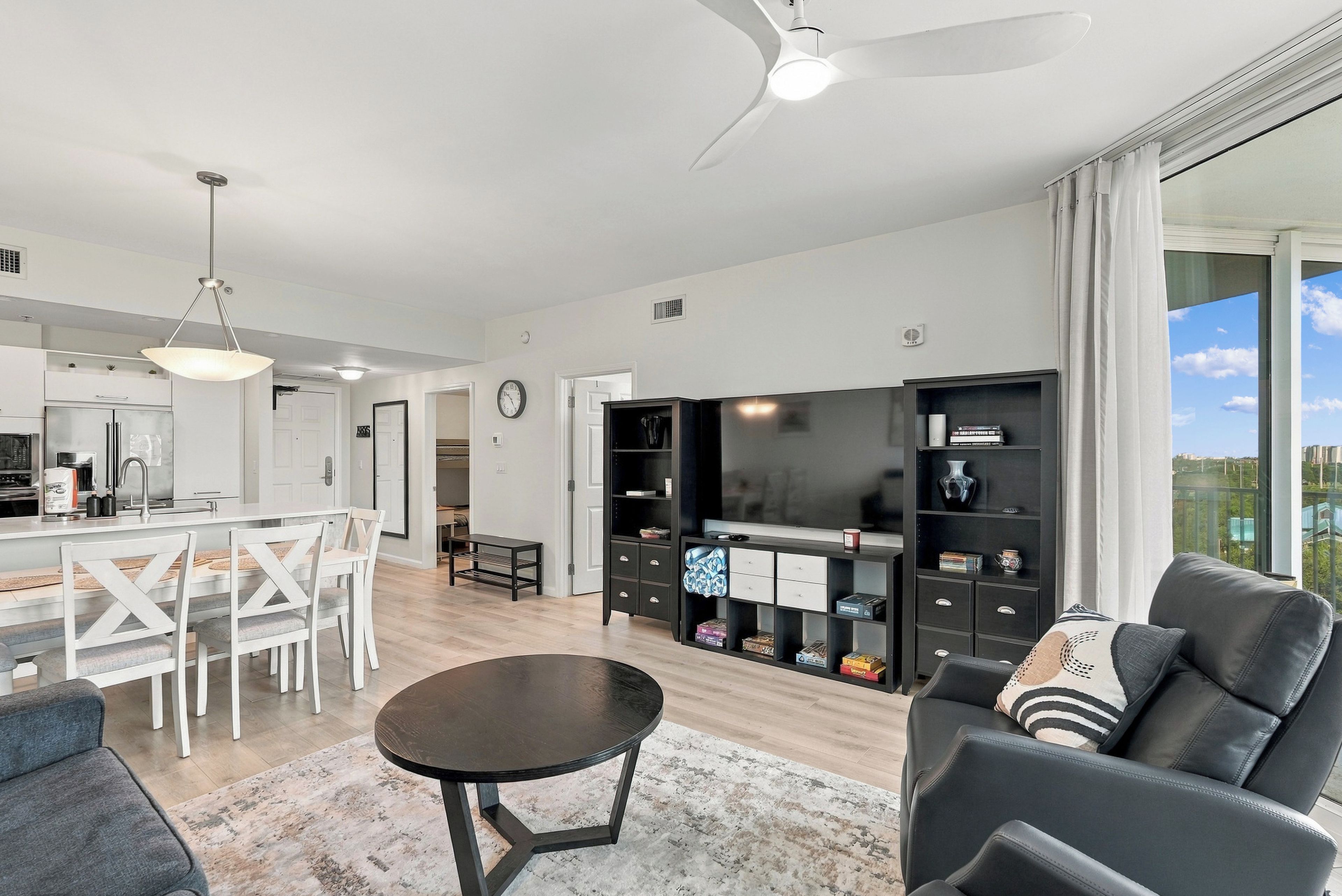 A modern living room with a black sofa, a dining area with a white table and chairs, and a kitchen with a refrigerator and a coffee machine.