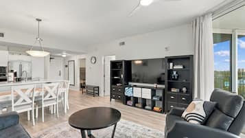 A modern living room with a black sofa, a dining area with a white table and chairs, and a kitchen with a refrigerator and a coffee machine.