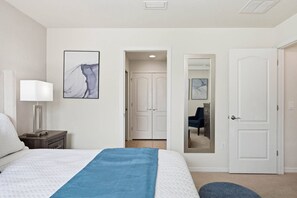 Ein Hotelzimmer mit Bett, Nachttischlampe, blauer Tagesdecke, einem abstrakten Gemälde, einer Tür und einem Spiegel, der einen blauen Sessel und eine Kommode reflektiert.
