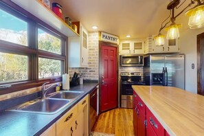 Una cocina con encimera de madera, electrodomésticos de acero inoxidable, una puerta de despensa roja y un ventanal con vistas a árboles.