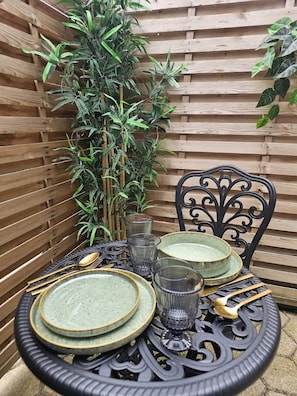 A round table with plates, glasses, and cutlery, set against a wooden backdrop with a bamboo plant.