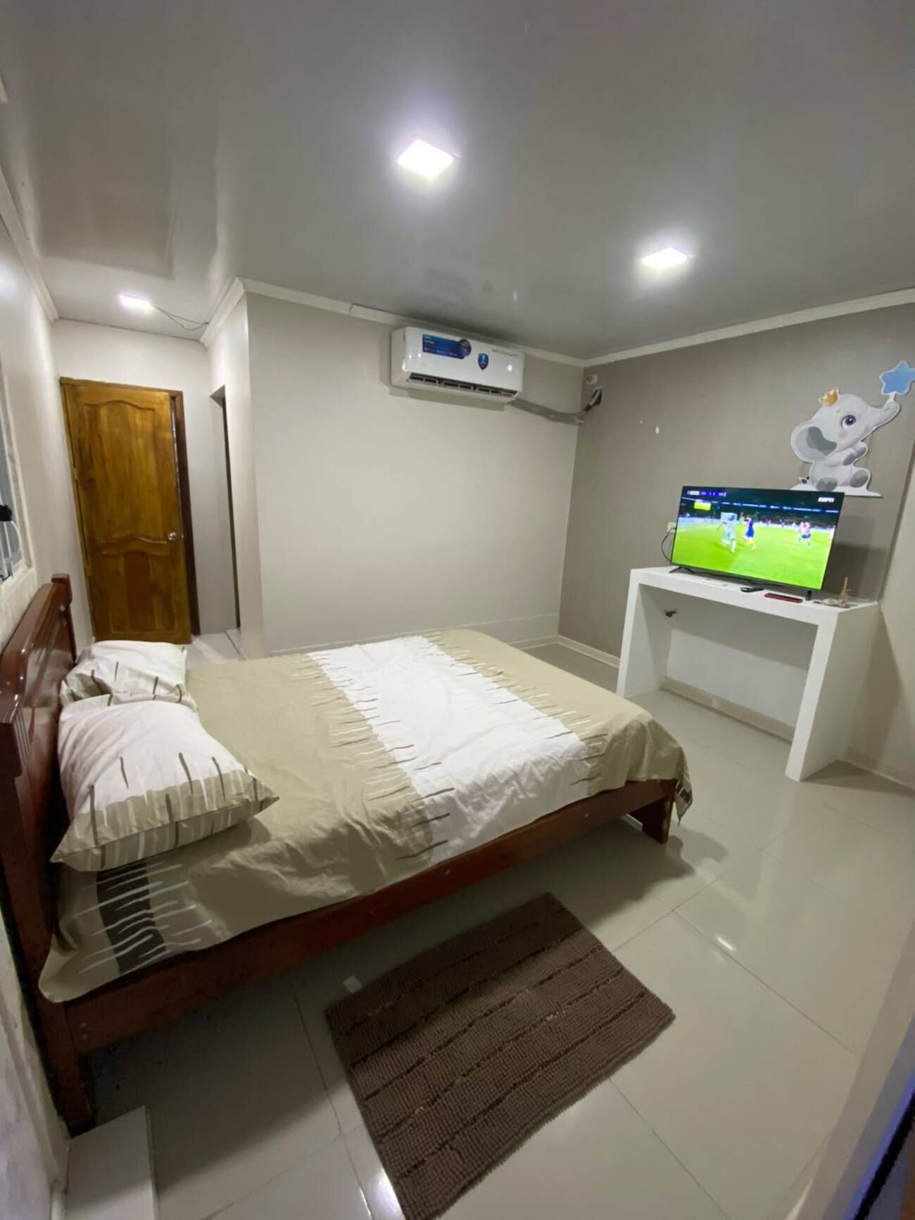A bedroom with a bed, a television on a stand, an air conditioning unit, and a door.