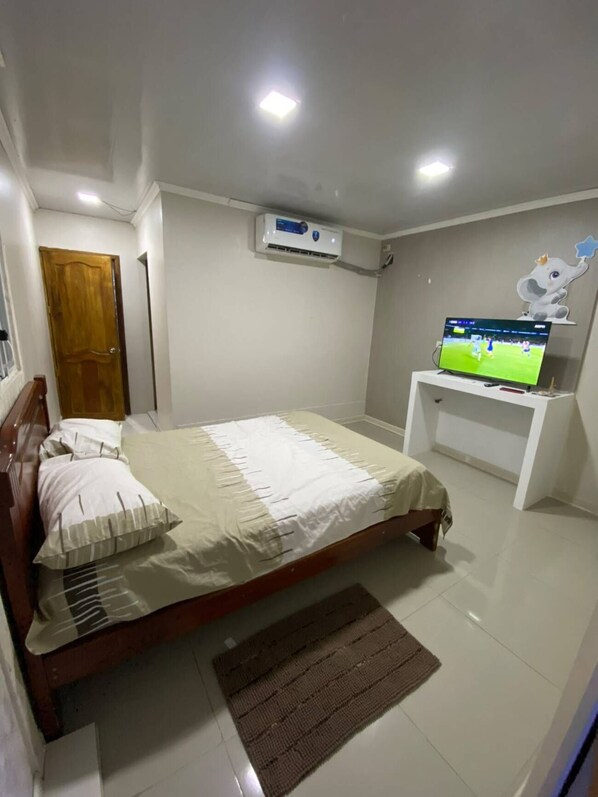 A bedroom with a bed, a television on a stand, an air conditioning unit, and a door.
