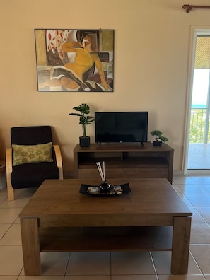 A living room with a wooden coffee table, a black sofa with a green pillow, a flat-screen TV on a wooden stand, a painting on the wall, and a view of the outdoors through a glass door. - The sea view house of Xenia and Panos by MPS (Paralia Avlidos)