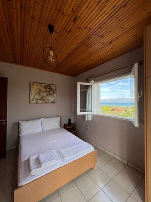 A bedroom with a wooden ceiling, a bed with white linens, a painting on the wall, and a window with a view of a landscape. - The sea view house of Xenia and Panos by MPS (Paralia Avlidos)