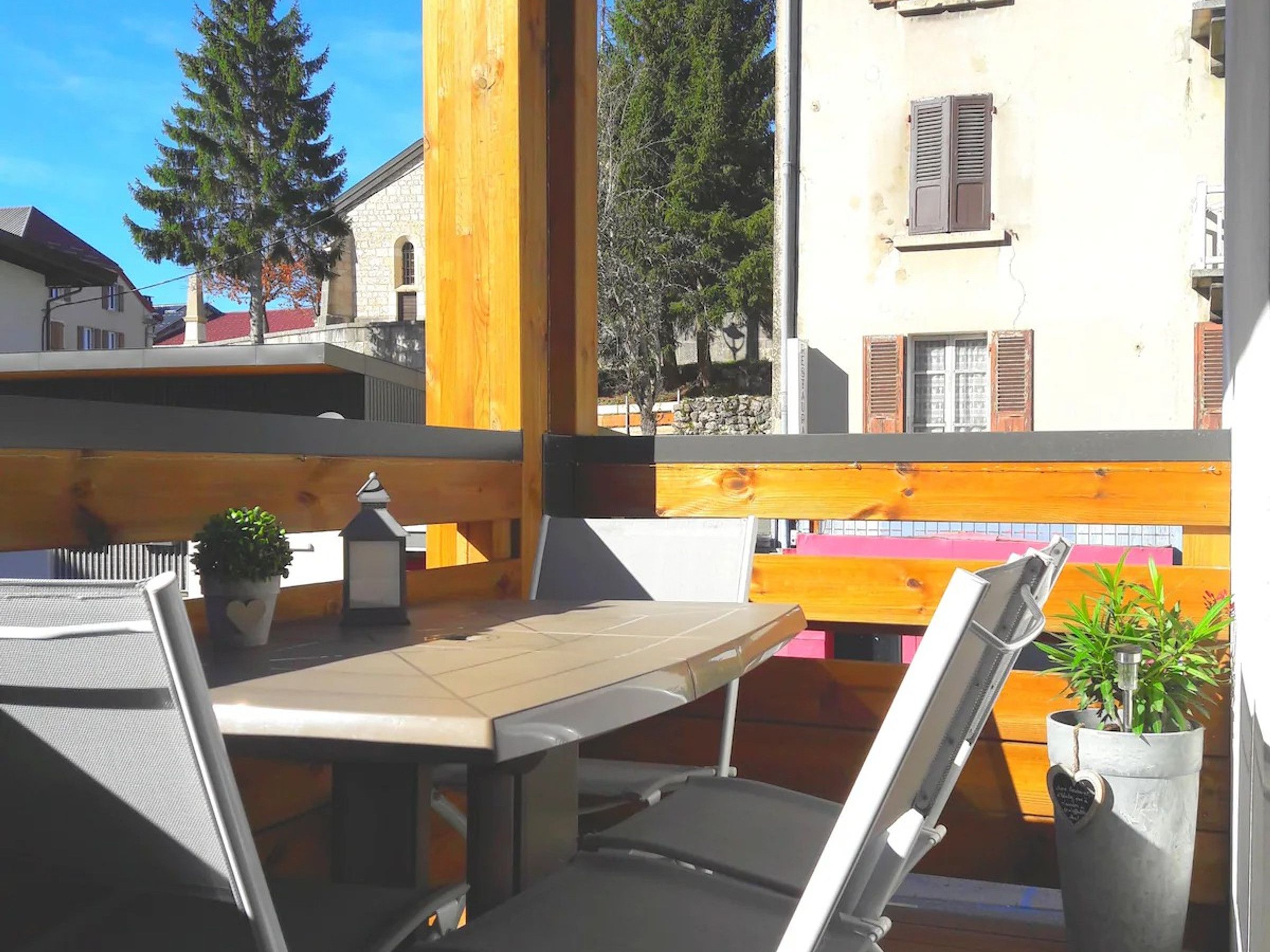 An outdoor seating area with a table and chairs, surrounded by a wooden fence, overlooking a residential street with houses and trees.
