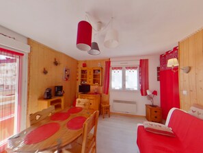 A cozy room with a wooden dining table, a red sofa, and a window with red curtains. - Central studio with balcony - 3 guests (Les Rousses)