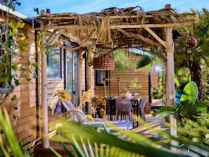 Una pergola in legno con tavolo e sedie sotto, circondata da vegetazione rigogliosa e un muro in mattoni.