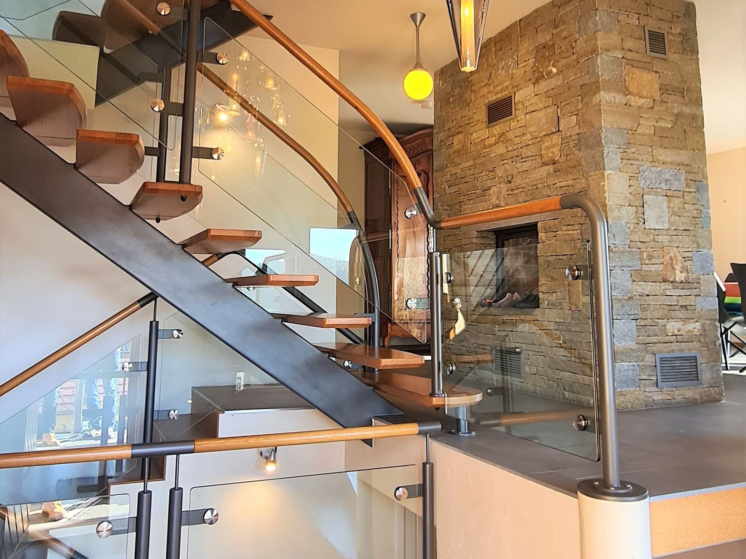 A modern interior with a glass staircase, a stone fireplace, and a hanging light fixture.