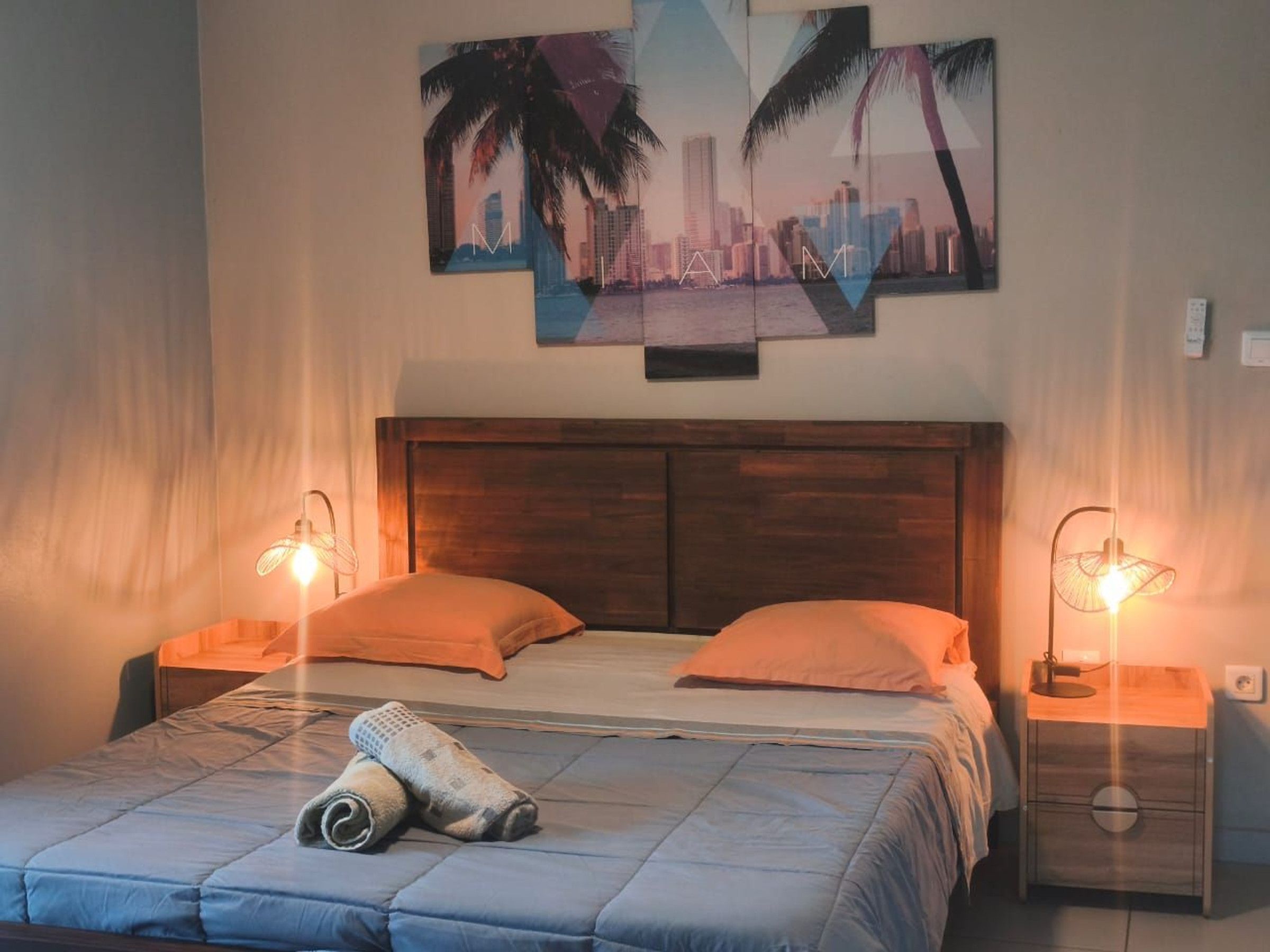 A bedroom with a wooden headboard, two bedside tables with lamps, a bed with a blue quilt, and a cityscape painting on the wall.