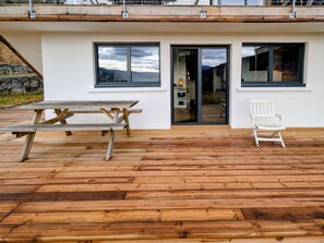 A wooden deck with a picnic table and a white chair, a white building with a balcony, and a glass door leading to a room. - Apartment with panoramic view - 4 guests (Cleurie)
