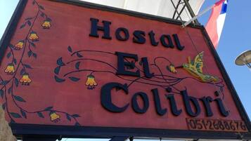 A sign for 'Hostal El Colibri' with a bird and floral design, and a contact number.
