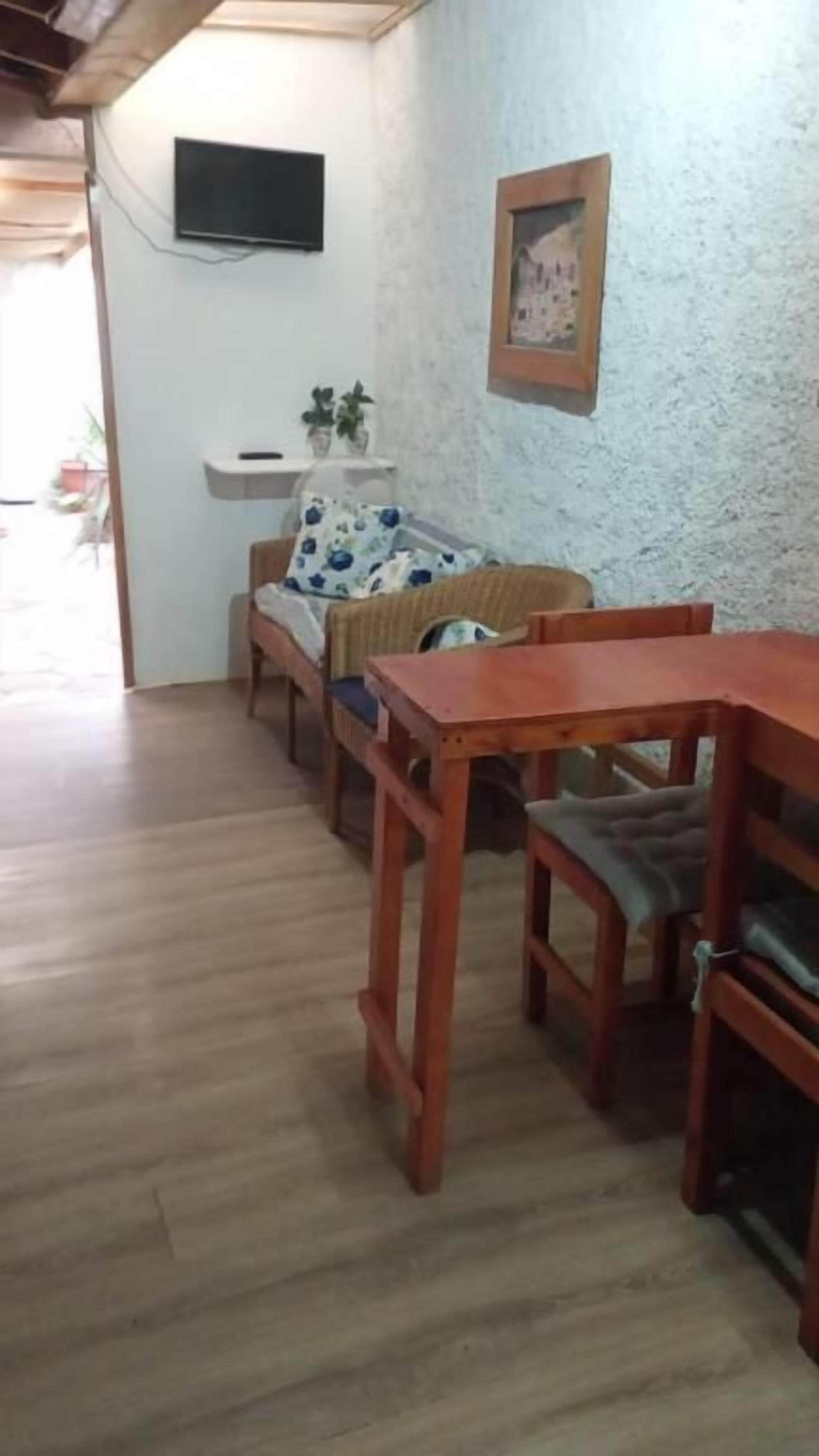 Una habitación con una mesa de comedor de madera, un sofá con un cojín floral y un televisor montado en la pared.