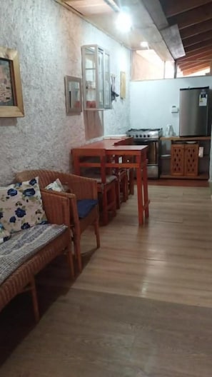 Zona d'estar amb un sofà, taula de dinar de fusta i cadires. Es veu una cuina amb nevera i fogó al fons.