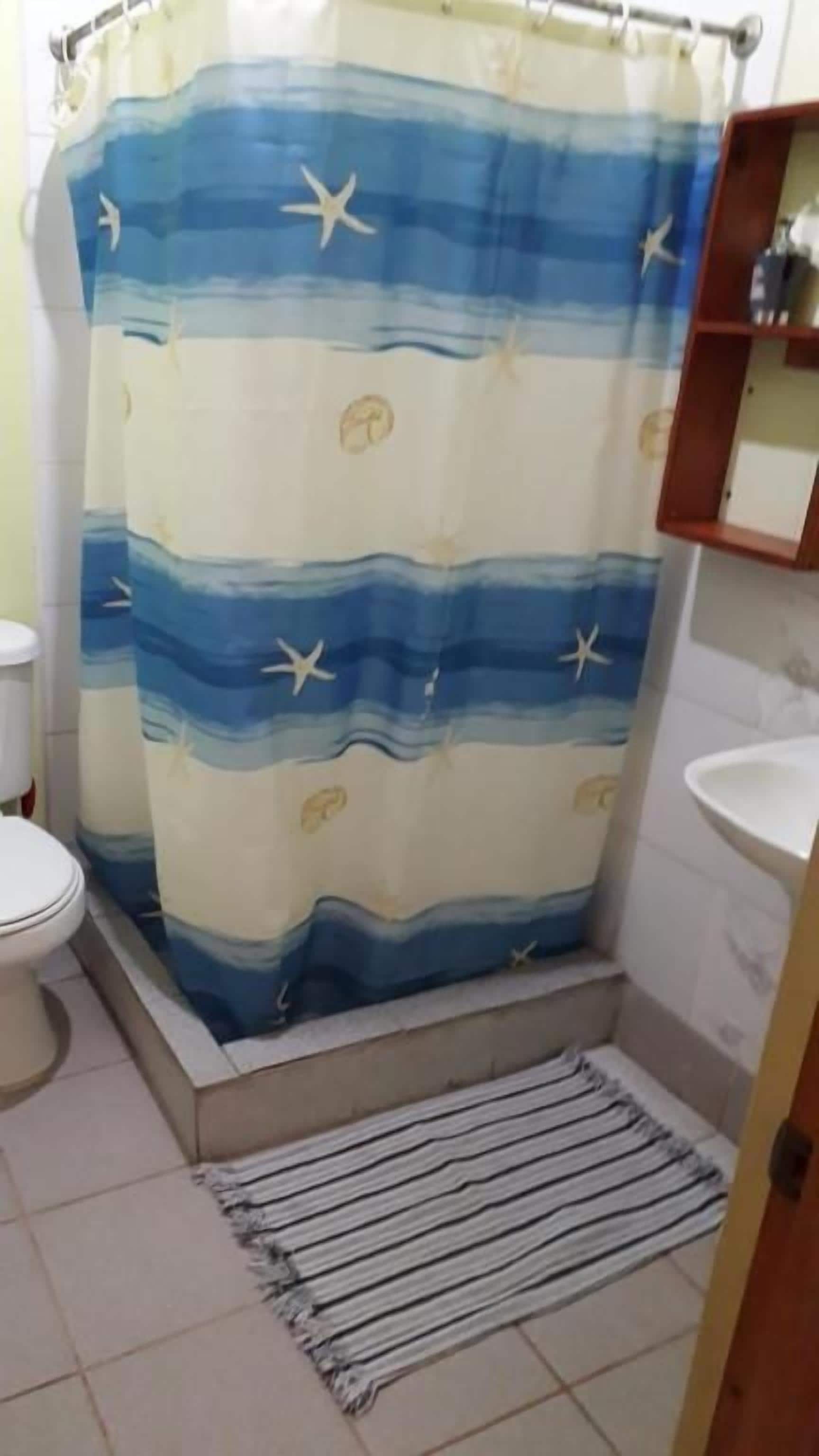 Un baño con cortina de ducha, inodoro, lavamanos y un estante pequeño.