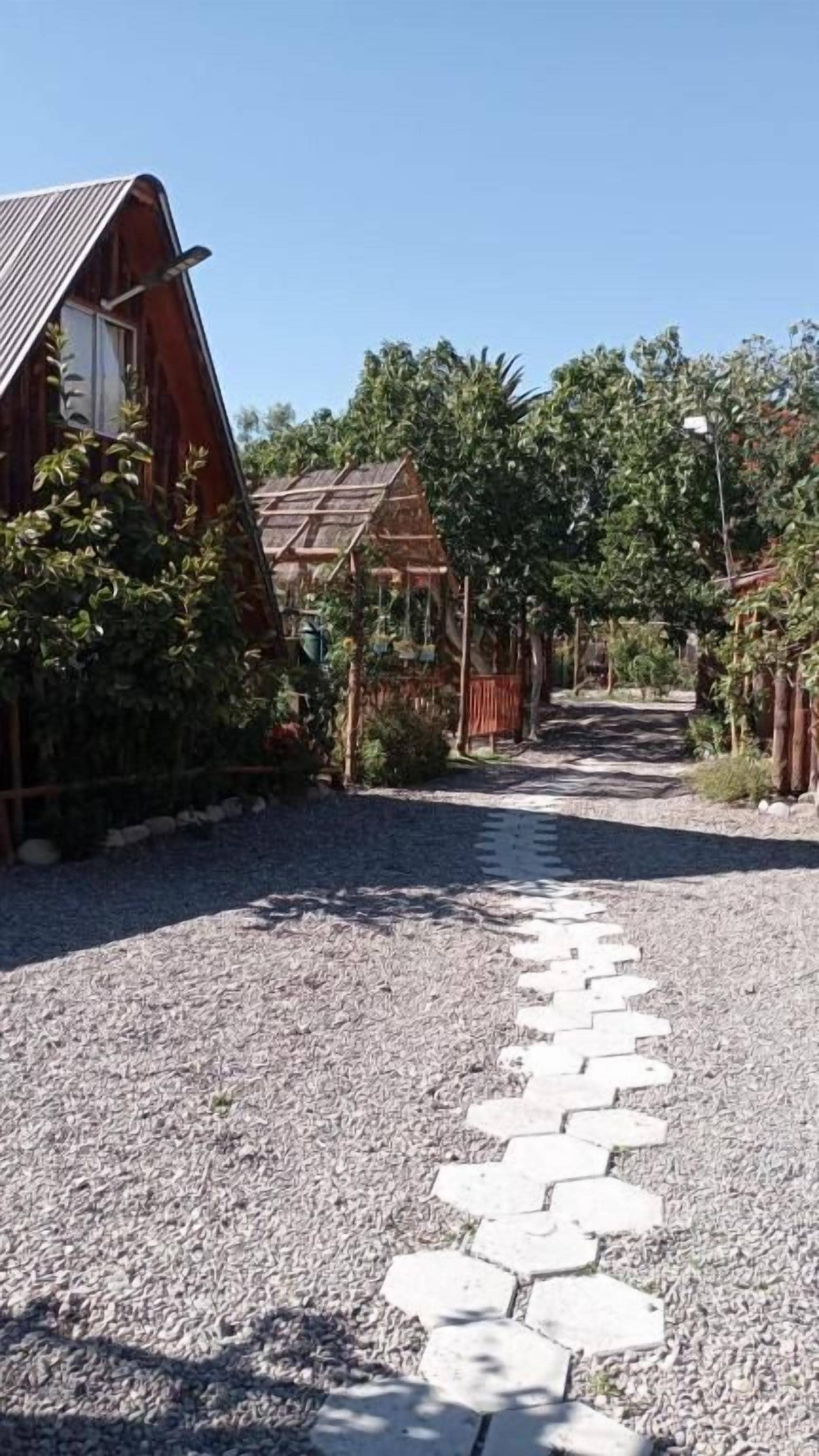 A gravel path leading to a wooden building with a sloped roof, surrounded by greenery and a clear sky.