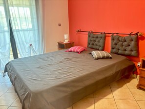 A bedroom with a bed, pillows, a nightstand, a lamp, and a window with curtains. - 3-Bedroom Air-conditioned Apartment near the Beach (Petite Île)