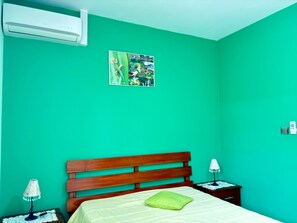 Une chambre avec un lit simple, une tête de lit en bois, un mur vert, une petite table de chevet avec une lampe et un climatiseur fixé au mur.