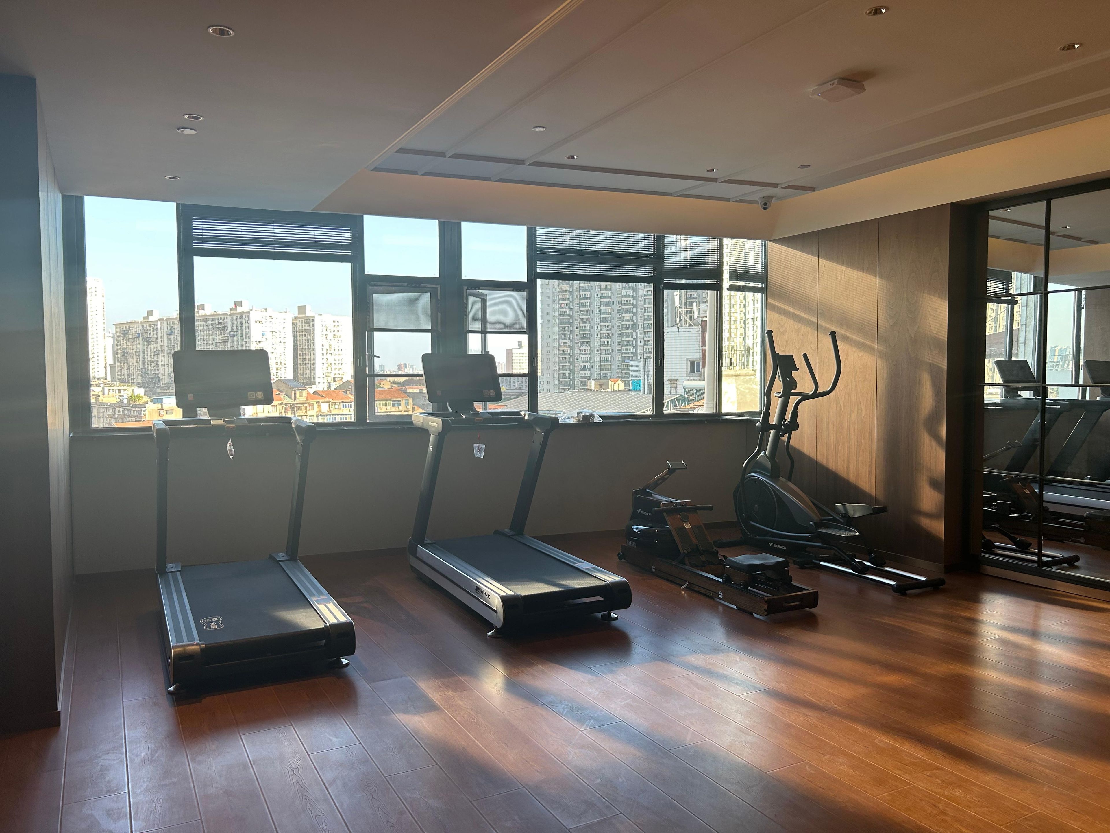 Academia com esteiras, elíptica e equipamentos de exercícios. Grandes janelas oferecem vista para o horizonte da cidade.