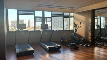 Academia com esteiras, elíptica e equipamentos de exercícios. Grandes janelas oferecem vista para o horizonte da cidade.