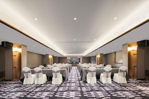 Uma sala de conferências de hotel, com fileiras de mesas e cadeiras, iluminação no teto e um tapete estampado.