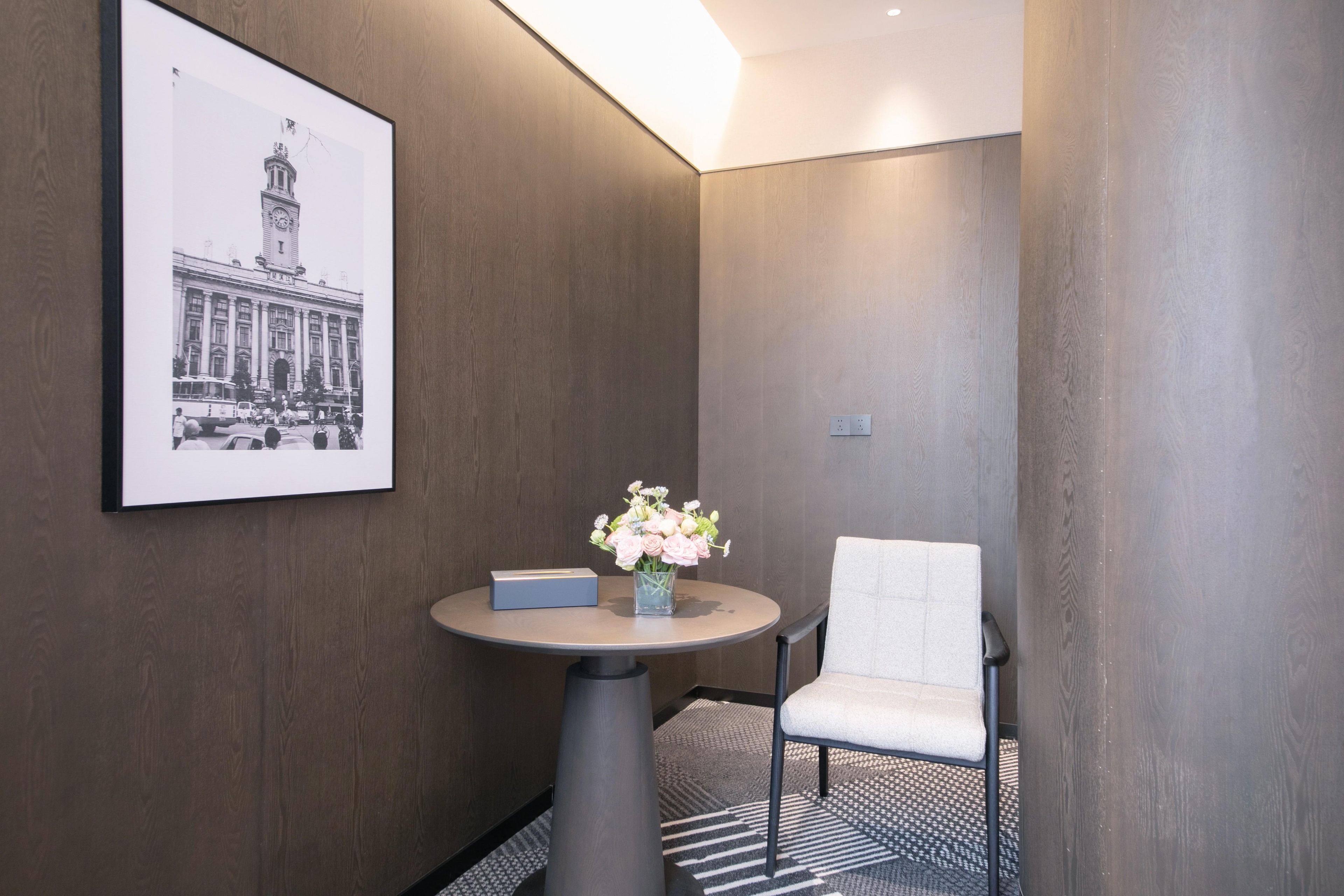 Een hotelkamer met een ingelijste zwart-witfoto aan de muur, een klein rond tafeltje met een bloemenvaas, een stoel en een vloerbedekking.