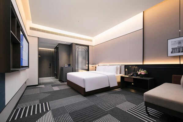 A modern hotel room with a large bed, a desk, a chair, a nightstand, a lamp, and a framed picture on the wall.