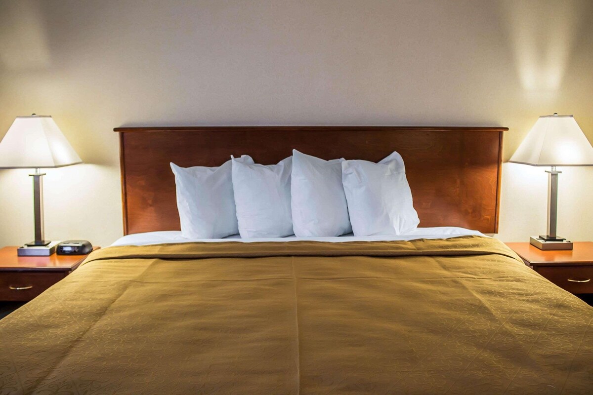 Una cama bien hecha con una colcha marrón y almohadas blancas, flanqueada por dos mesitas de noche con lámparas.