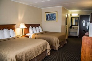 Ein Hotelzimmer mit zwei Betten, einem Nachttisch mit Lampe, einem Bild an der Wand, einem Badezimmer mit Waschbecken und Spiegel sowie einem Holztisch.