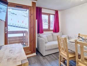 Een gezellige kamer met een bank, een eettafel met stoelen, een raam met gordijnen en uitzicht op een besneeerd landschap.