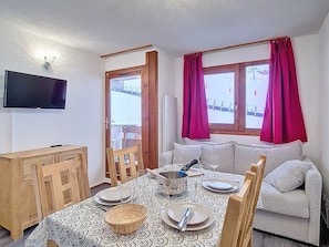 Ein Essbereich mit einem Tisch für zwei Personen, einer Couch, einem an der Wand befestigten Fernseher und einem Fenster mit Vorhängen.
