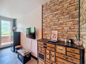 A room with a brick wall, a television mounted on the wall, a wooden chest of drawers, and a window with a view of greenery. - Bright apartment, balcony - 4 guests (Les Menuires)