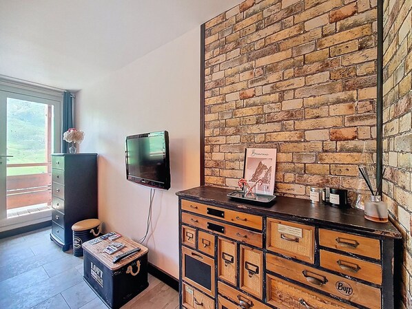 Una stanza con parete in mattoni, una televisione fissata al muro, un comò in legno e una finestra con vista sul verde.