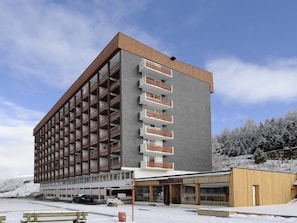 A multi-story hotel with a modern design, featuring a combination of brick and wood exteriors, and a parking area in front. - Bright apartment, balcony - 4 guests (Les Menuires)