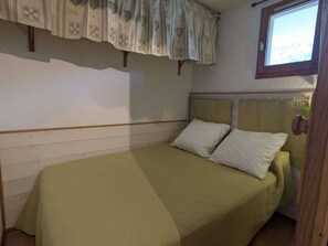 Ein Einzelbett mit grüner Bettdecke und zwei Kissen, ein Fenster mit Holzrahmen und einem gemusterten Vorhang.