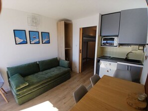 A compact living space with a green sofa, a wooden dining table, and a kitchenette. - 2-room apartment - 5 guests, south balcony, L'Alpe d'Huez (Huez)